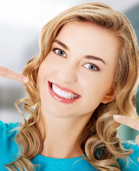 Woman with blond hair in teal shirt pointing to her perfect smile with both hands