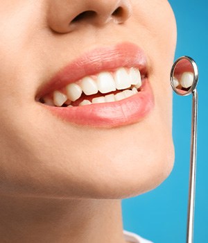 Nose-to-neck closeup of woman smiling holding dental mirror to teeth with blue background